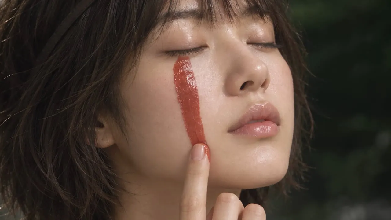 Uma mulher de olhos fechados traça uma linha de creme vermelho pela bochecha.